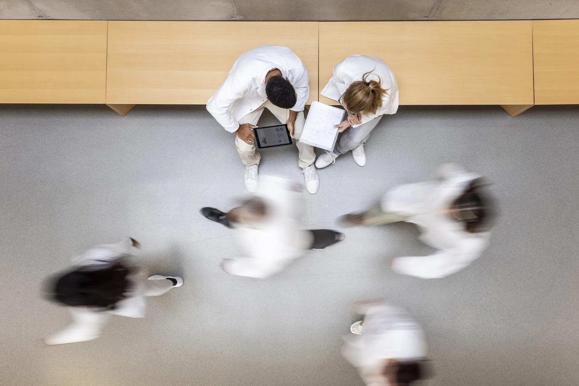 Deux professionnels en blouse blanche assis dans un couloir, entourés de personnes en mouvement.